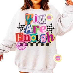 Pu&ograve; includere: Una felpa grigio chiaro con le parole "You Are Enough" in lettere colorate ritagliate. Il design include stelle a scacchi e un bordo a scacchi, con un timbro rosa "Original".