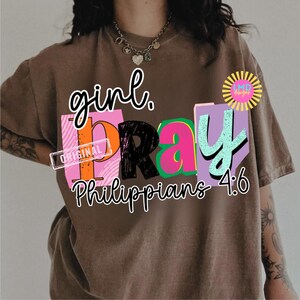 May include: Brown t-shirt with the words "girl. PRAY" in a colorful, block letter design. The text "Philippians 4:6" is below the word "PRAY". The shirt has a vintage, worn look.
