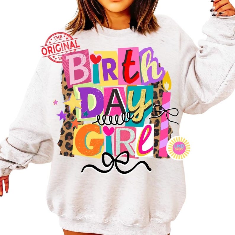 May include: A light gray sweatshirt with a colorful "Birthday Girl" graphic. The design includes the words "Birthday Girl" in various colors, leopard print accents, stars, candles, and bows. The text "The Original" is stamped in red.