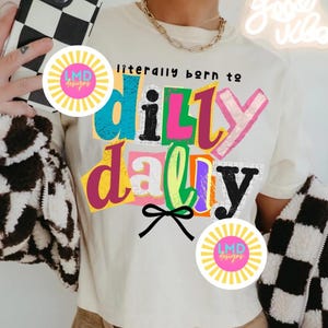 May include: Cream-colored t-shirt with the phrase "literally born to dilly dally" in colorful, block letters. The shirt features a black bow graphic and a gold chain necklace. A black and white checkered jacket is visible.