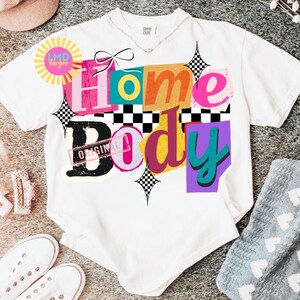 May include: White t-shirt with the words "Home Body" in colorful, layered text. The design includes a bow, checkerboard accents, and the word "Original" stamped on the "Body" text. The shirt has a crew neck and short sleeves.