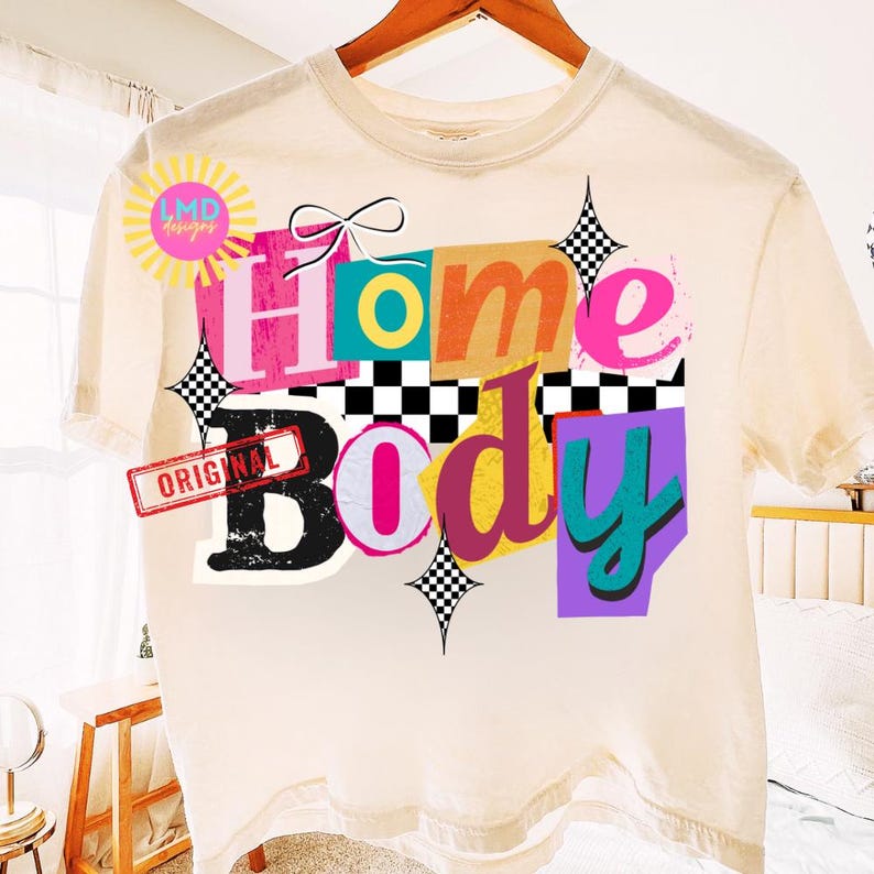 May include: Cream-colored t-shirt with a colorful graphic design that reads "Home Body." The design features the words in various colors and fonts, with a bow, stars, and a checkerboard pattern. The word "ORIGINAL" is stamped on the shirt.