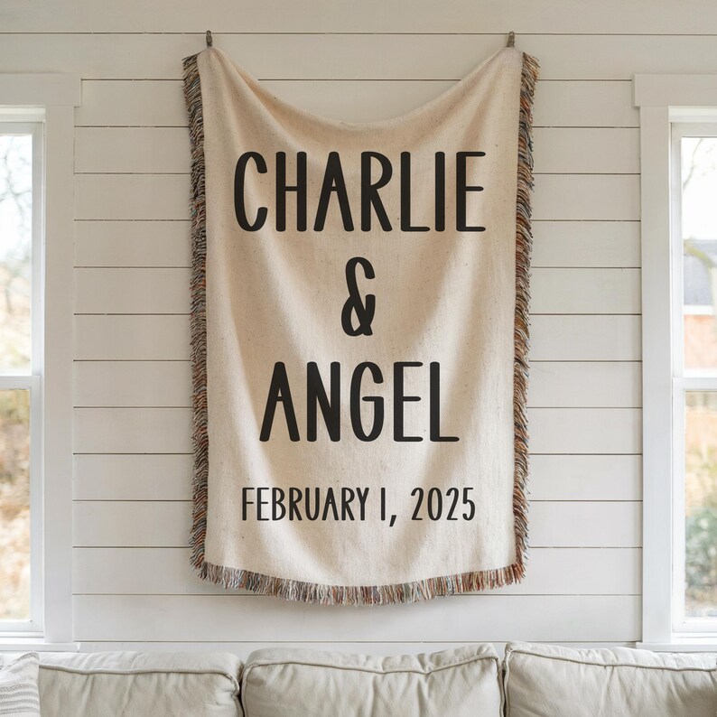 May include: A cream-colored woven throw blanket with fringe on all sides. The blanket has black text that reads "Charlie & Angel February 1, 2025".
