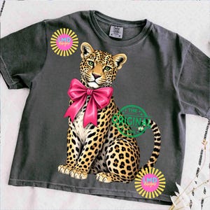 May include: Dark gray t-shirt featuring a leopard with a pink bow. The leopard has a spotted coat and green eyes. The shirt also has the text "THE ORIGINAL" and "LMD designs" in a circular design.