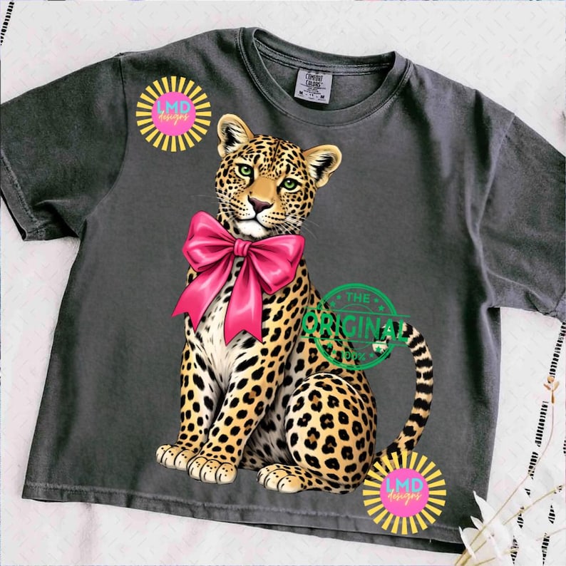 May include: Dark gray t-shirt featuring a leopard with a pink bow. The leopard has a spotted coat and green eyes. The shirt also has the text "THE ORIGINAL" and "LMD designs" in a circular design.