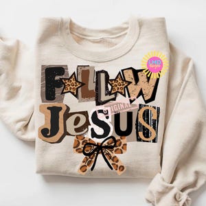 K&ouml;nnte beinhalten: Beiges Sweatshirt mit dem Schriftzug "FOLLOW JESUS" in einem Collage-Design. Die Buchstaben sind in verschiedenen Schriftarten und Farben gehalten, darunter Braun, Schwarz und Leopardenmuster. Eine Schleife mit Leopardenmuster befindet sich unten.