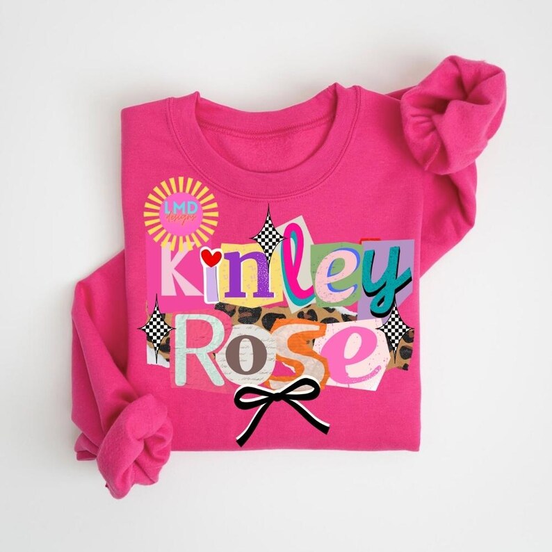 May include: A bright pink sweatshirt with the name "Kinley Rose" in a collage style design. The design includes various colors, patterns, and a black bow. The sweatshirt also features the text "LMD designs" in a yellow circle.