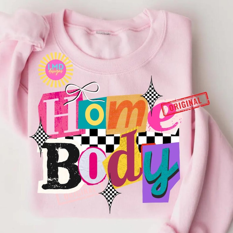 May include: A light pink sweatshirt with the words "Home Body" in a colorful, collage-style design. The design includes a bow, checkered patterns, and the word "ORIGINAL" stamped on the right side. A sun-shaped logo is on the left side.