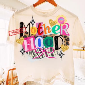 May include: Cream-colored t-shirt with the word "Motherhood" in a colorful, collage-style design. The design includes leopard print, zebra stripes, glitter hearts, and a pink bow. The word "ORIGINAL" is stamped on the shirt.