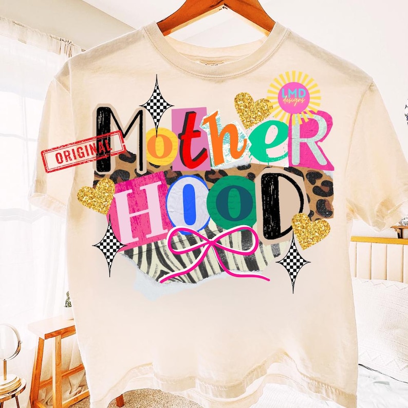 May include: Cream-colored t-shirt with the word "Motherhood" in a colorful, collage-style design. The design includes leopard print, zebra stripes, glitter hearts, and a pink bow. The word "ORIGINAL" is stamped on the shirt.