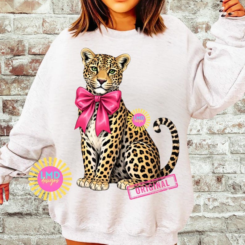 May include: White sweatshirt featuring a leopard with a pink bow. The leopard has a spotted coat and green eyes. The design includes the text "ORIGINAL" on a pink rectangle and a sunburst logo with the text "LMD designs".