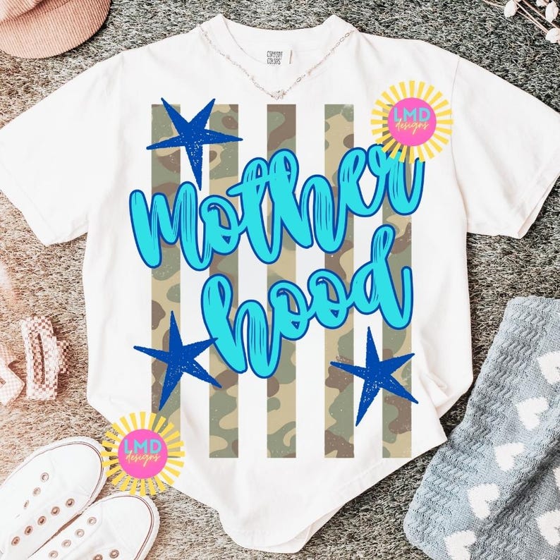 May include: White t-shirt with the word "Motherhood" in blue script, accented with blue stars and a camouflage stripe design. The shirt also features a sunburst logo with the text "LMD designs".