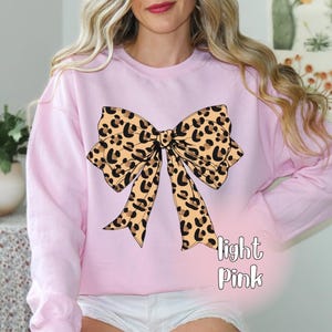 May include: Light pink sweatshirt featuring a large leopard print bow design. The bow has a tan background with black spots. The words "light Pink" are printed in white script below the bow. The sweatshirt has long sleeves.