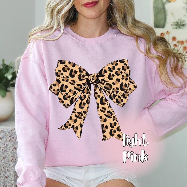 May include: Light pink sweatshirt featuring a large leopard print bow design. The bow has a tan background with black spots. The words "light Pink" are printed in white script below the bow. The sweatshirt has long sleeves.