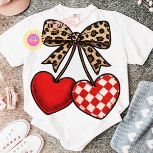 May include: White t-shirt featuring a large graphic of two cherries. One cherry is solid red, the other is red and white checkered. A leopard print bow sits above the cherries. The shirt has the text "Original" and "LMD designs".