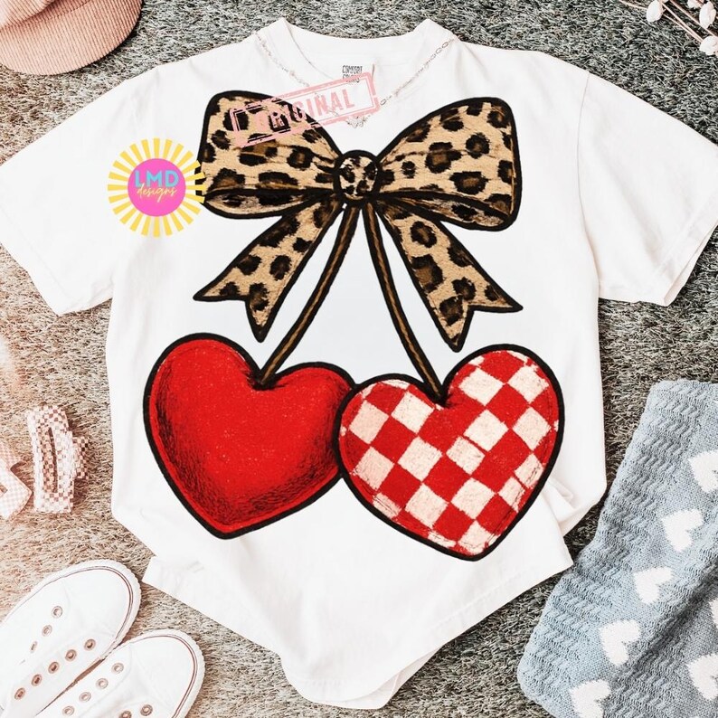 May include: White t-shirt featuring a large graphic of two cherries. One cherry is solid red, the other is red and white checkered. A leopard print bow sits above the cherries. The shirt has the text "Original" and "LMD designs".