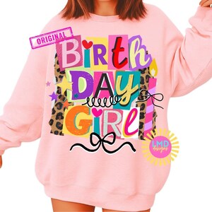 May include: A light pink sweatshirt with the words "BIRTHDAY GIRL" in a colorful, block letter design. The design includes hearts, stars, and a bow. The word "ORIGINAL" is in a pink rectangle in the upper left corner.