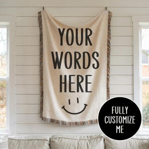 May include: A white woven tapestry with the text "YOUR WORDS HERE" and a smiley face. The tapestry has a fringe border and is hanging on a white wall. The text "FULLY CUSTOMIZE ME" is in a black circle below the tapestry.