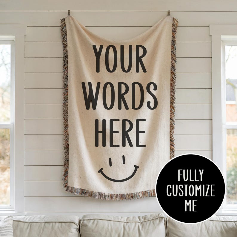 May include: A white woven tapestry with the text "YOUR WORDS HERE" and a smiley face. The tapestry has a fringe border and is hanging on a white wall. The text "FULLY CUSTOMIZE ME" is in a black circle below the tapestry.