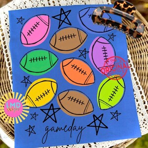 May include: A blue t-shirt featuring a repeating pattern of colorful footballs in pink, brown, green, orange, and yellow, interspersed with black hand-drawn stars and the word gameday written in a cursive black script at the bottom.