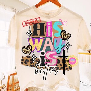 May include: Cream-colored t-shirt with a colorful graphic design. The design includes the words "His Way is better" in various fonts and colors, along with a cross, hearts, and stars. The word "Original" is in a red rectangle.