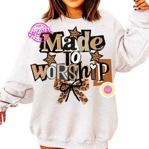 K&ouml;nnte beinhalten: Ein hellgraues Sweatshirt mit dem Schriftzug "Made to Worship" in verschiedenen Schriftarten und Farben. Das Design umfasst Sterne und Schleifen mit Leopardenmuster. Ein rosa Stempel mit der Aufschrift "The Original" ist oben links zu sehen.
