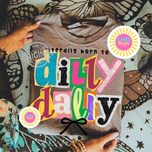 May include: A taupe t-shirt with the phrase "literally born to dilly dally" in colorful, cut-out style letters. The design includes a black bow and the LMD designs logo. The shirt is held by hands.