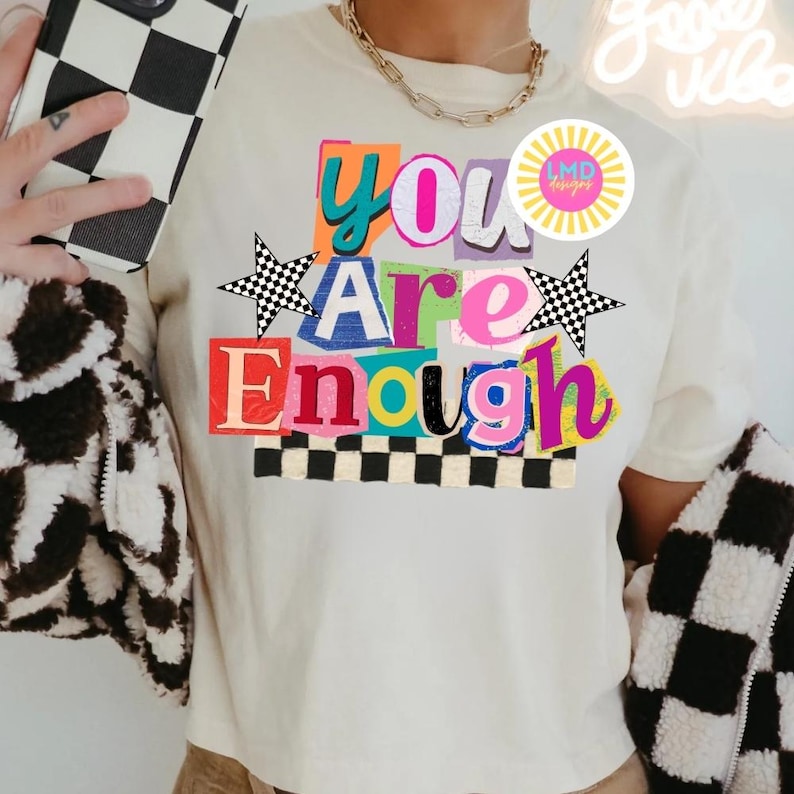 Pu&ograve; includere: T-shirt color crema con le parole "You Are Enough" in lettere colorate in stile ritagliato. La maglietta presenta stelle a scacchi bianche e nere e un bordo a scacchi nella parte inferiore. &Egrave; visibile una collana a catena d'oro.