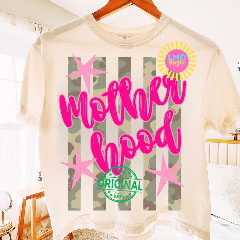 May include: Cream-colored t-shirt with the word "Motherhood" in bright pink script. The design includes pink stars, vertical stripes with a camouflage pattern, and a "100% Original" seal. A yellow sun with the text "LMD designs" is also present.