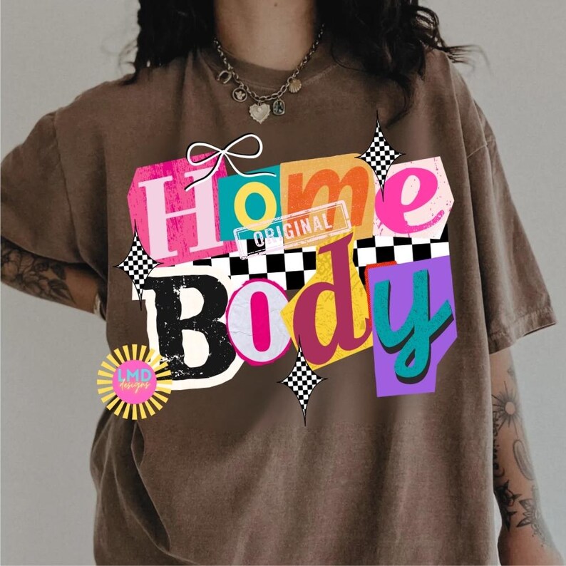 May include: Brown t-shirt with a colorful graphic that says "Home Body." The design features the words in various colors and fonts, with a bow and checkered accents. The shirt also has a small logo that says "LMD designs."