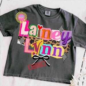 May include: Dark grey t-shirt with the name "Lainey Lynn" in colorful, layered text. The design includes a heart, leopard print, and a black and white bow. The word "ORIGINAL" is stamped on the shirt.