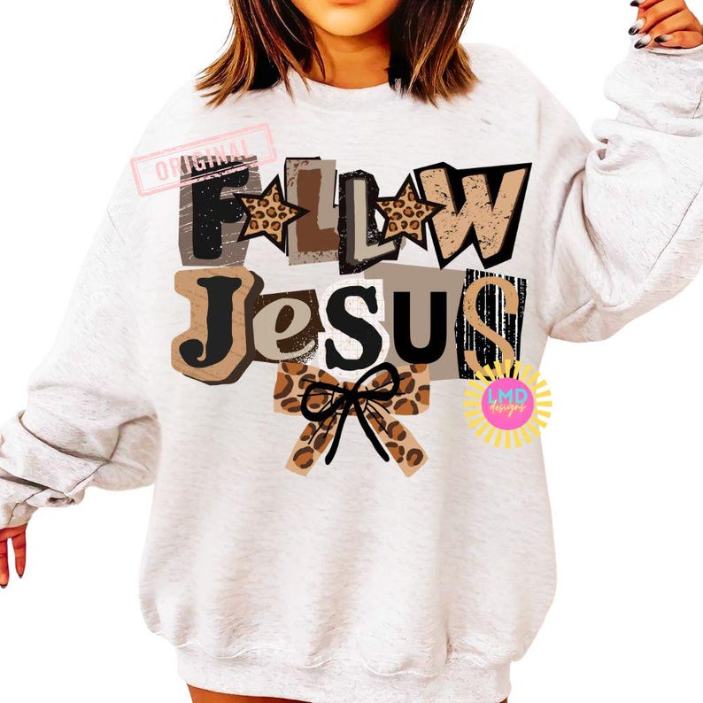 K&ouml;nnte beinhalten: Ein hellgraues Sweatshirt mit den Worten "FOLLOW JESUS" in einem mehrschichtigen Design. Die Buchstaben haben verschiedene Texturen, darunter Leopardenmuster und Braunt&ouml;ne. Eine Schleife mit Leopardenmuster befindet sich unten. Das Wort "ORIGINAL" ist oben links gestempelt.