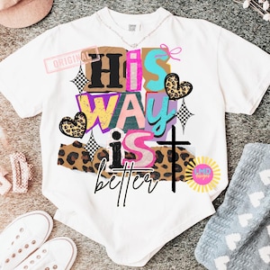 May include: White t-shirt with the phrase "His Way is Better" in a colorful, layered design. The design includes a cross, hearts, and checkered elements. The shirt has a crew neck and short sleeves.