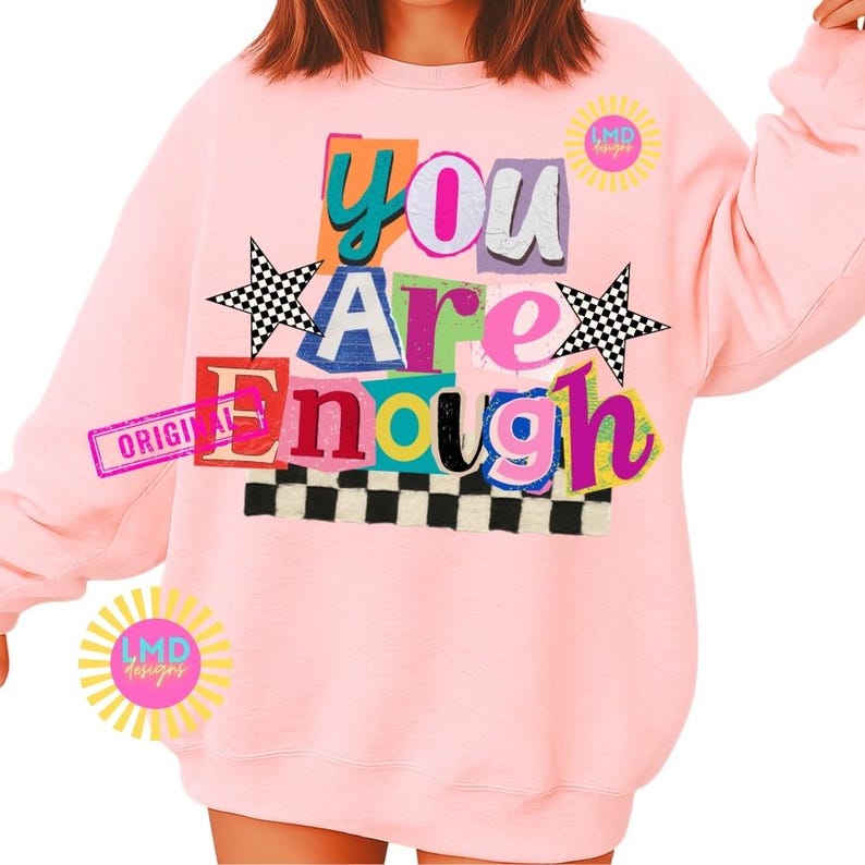 Pu&ograve; includere: Una felpa rosa chiaro con la scritta "You Are Enough" in un design colorato in stile collage. Il design include stelle a scacchi bianche e nere e un bordo a scacchi bianchi e neri nella parte inferiore. La felpa ha anche il testo "ORIGINAL" su un rettangolo rosa.