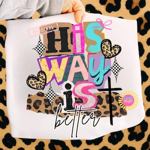 May include: White graphic tee with the text "HIS WAY IS better" in a colorful, layered design. The design includes a cross, hearts, and a leopard print pattern. The word "ORIGINAL" is stamped in pink.