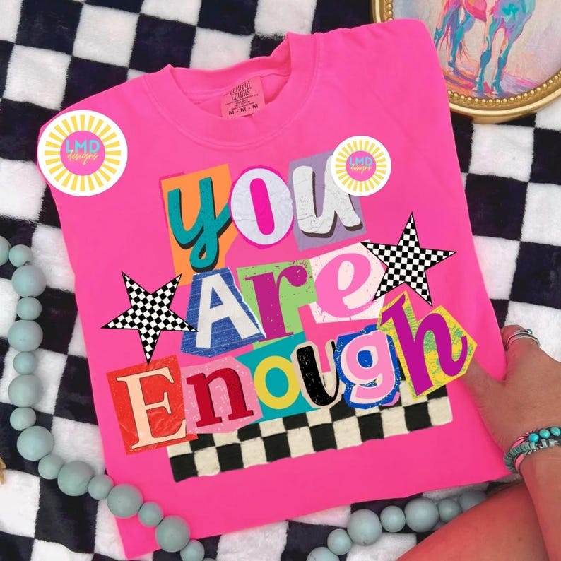 Pu&ograve; includere: Maglietta rosa acceso con la scritta "YOU ARE ENOUGH" in un design colorato in stile collage. Presenta due stelle a scacchi in bianco e nero e due adesivi "LMD designs". La maglietta &egrave; esposta su una superficie a scacchi in bianco e nero.
