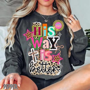 May include: Dark gray sweatshirt with the text "His Way is Better" in a colorful, layered design. The design includes leopard print stars, a cross, and a sunburst graphic. The sweatshirt has long sleeves.