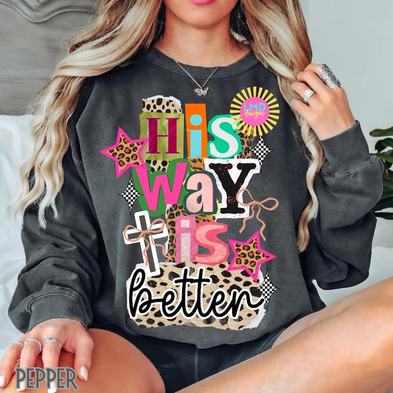 May include: Dark gray sweatshirt with the text "His Way is Better" in a colorful, layered design. The design includes leopard print stars, a cross, and a sunburst graphic. The sweatshirt has long sleeves.