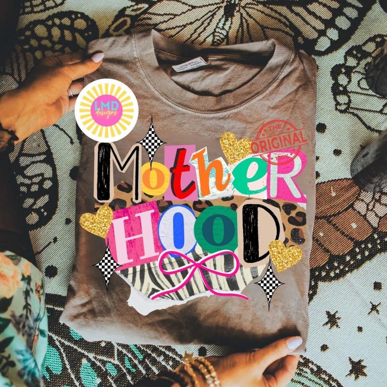 May include: A taupe-colored t-shirt with the word "Motherhood" in a collage style design. The design includes various patterns, including leopard print, zebra stripes, and gold glitter hearts. The text "The Original" is stamped on the top right.