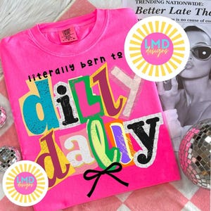 May include: A bright pink sweatshirt with the phrase "literally born to dilly dally" in a colorful, collage-style design. The shirt features a crew neck and long sleeves. The image includes the "LMD designs" logo and a black bow.