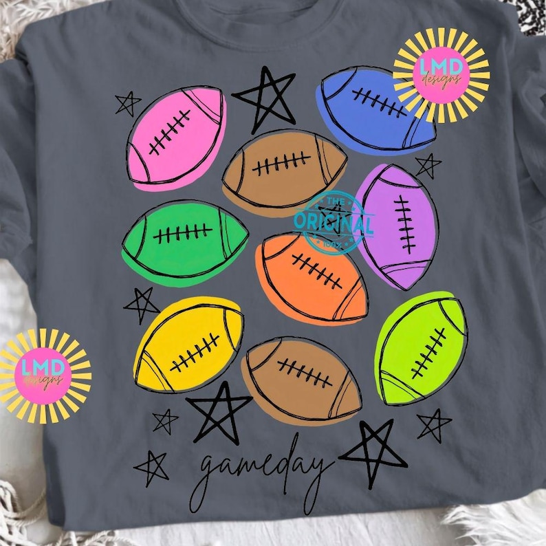 May include: A dark gray t-shirt featuring a colorful graphic print of multiple footballs in pink, brown, blue, purple, green, orange, yellow, and lime green, interspersed with black hand-drawn stars and the word gameday in cursive script.