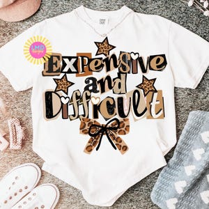 Pu&ograve; includere: T-shirt bianco con la scritta "Expensive and Difficult" in un mix di font e colori, con accenti marroni, neri e stampa leopardata. Il design include stelle e un fiocco. La maglietta &egrave; esposta su una superficie neutra.