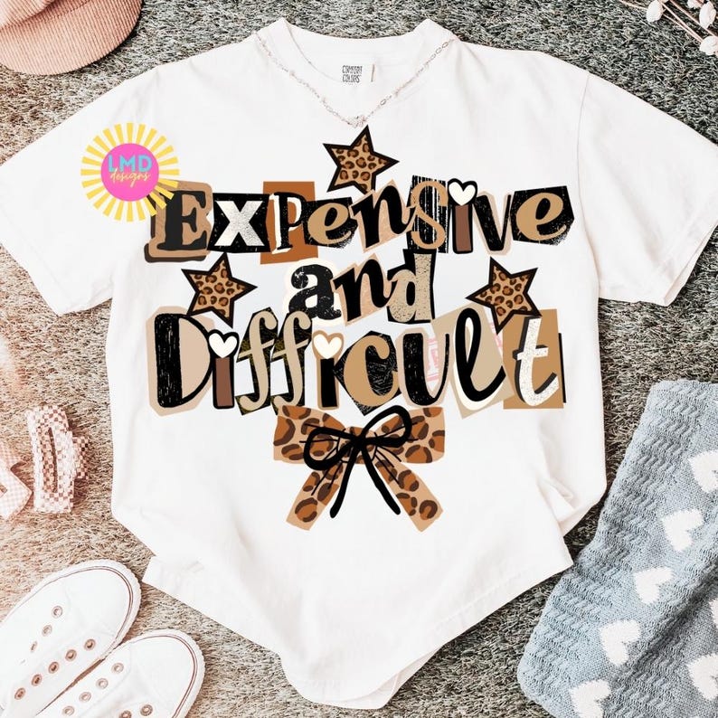 Pu&ograve; includere: T-shirt bianco con la scritta "Expensive and Difficult" in un mix di font e colori, con accenti marroni, neri e stampa leopardata. Il design include stelle e un fiocco. La maglietta &egrave; esposta su una superficie neutra.