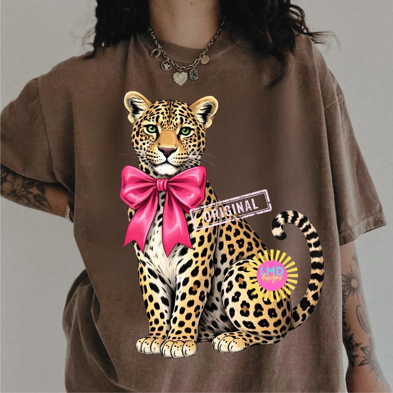 May include: Brown t-shirt featuring a seated leopard illustration with a large pink bow. The leopard has green eyes and a spotted coat. The word "ORIGINAL" is printed on a rectangular label.
