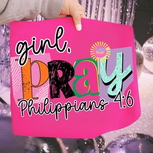 May include: A bright pink sign with the words "girl. PRAY Philippians 4:6" in various colorful fonts. The sign is held by a person with a light-colored sleeve. The background includes silver tinsel and disco balls.
