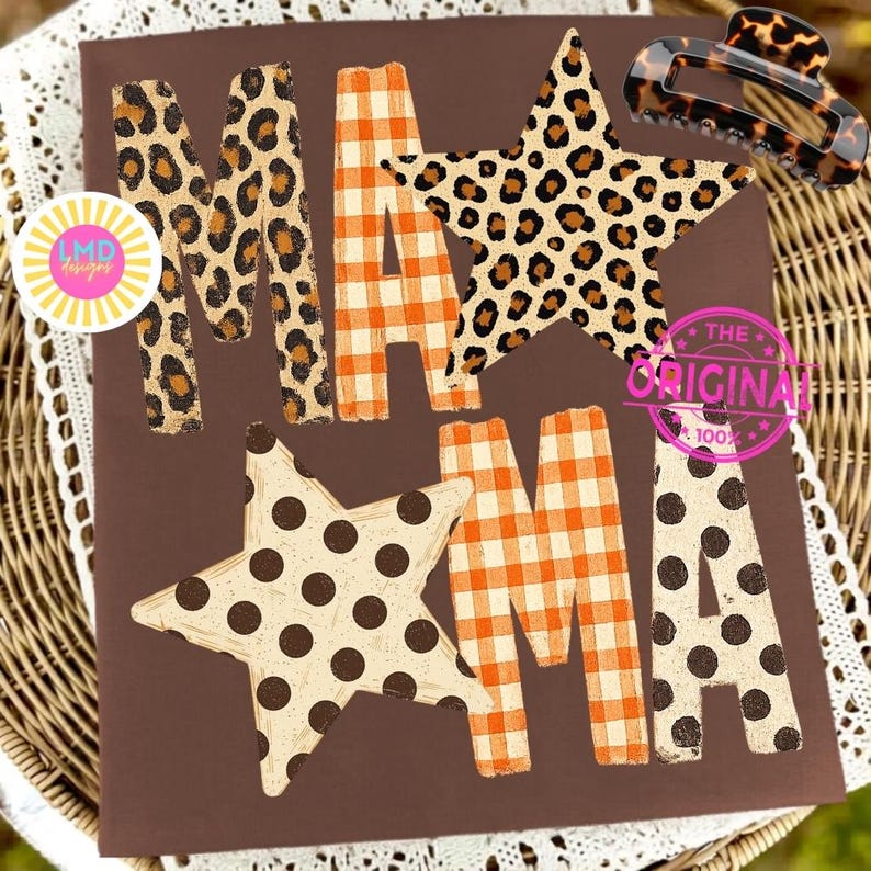 May include: A brown t-shirt featuring the text MAMA in a mix of patterns. The first M is leopard print, the A is orange and white gingham, the second M is a leopard print star, and the final A is orange gingham with a polka dot star overlay.