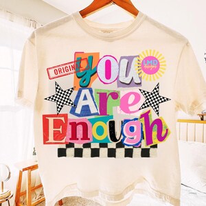 Pu&ograve; includere: T-shirt color crema con un design grafico colorato. Il design presenta le parole "You Are Enough" in stile collage, con un motivo a scacchiera e accenti a stella. La maglietta ha anche il testo "ORIGINAL" e "LMD designs".