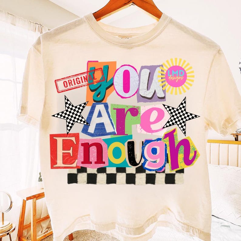 Pu&ograve; includere: T-shirt color crema con un design grafico colorato. Il design presenta le parole "You Are Enough" in stile collage, con un motivo a scacchiera e accenti a stella. La maglietta ha anche il testo "ORIGINAL" e "LMD designs".