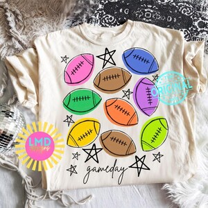 May include: A cream-colored t-shirt featuring a graphic print of multiple footballs in various colors including pink, blue, green, orange, yellow, and brown. The design includes black hand-drawn stars and the word gameday in cursive script.