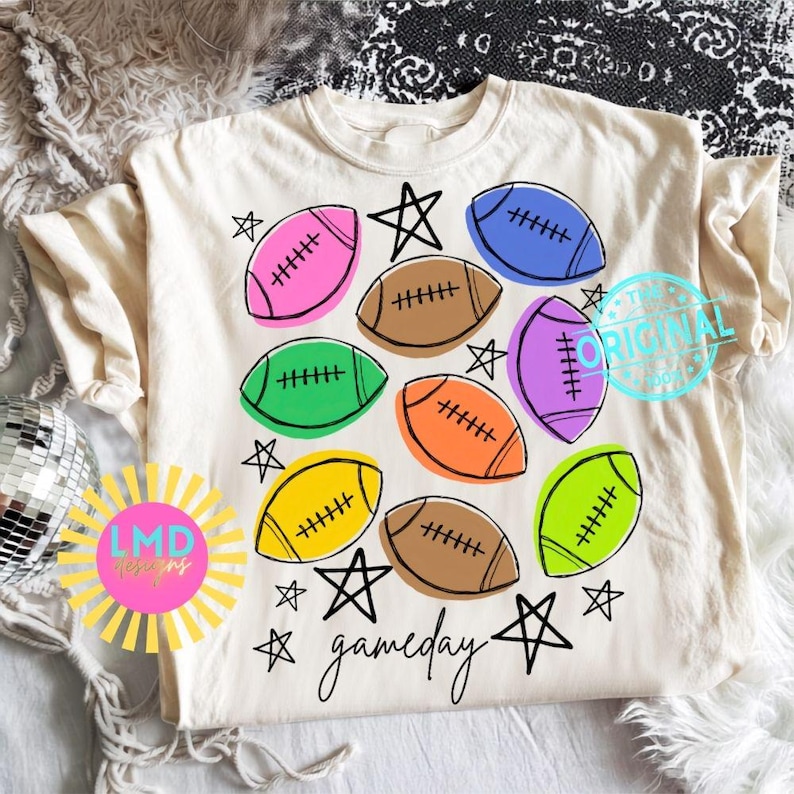 May include: A cream-colored t-shirt featuring a graphic print of multiple footballs in various colors including pink, blue, green, orange, yellow, and brown. The design includes black hand-drawn stars and the word gameday in cursive script.